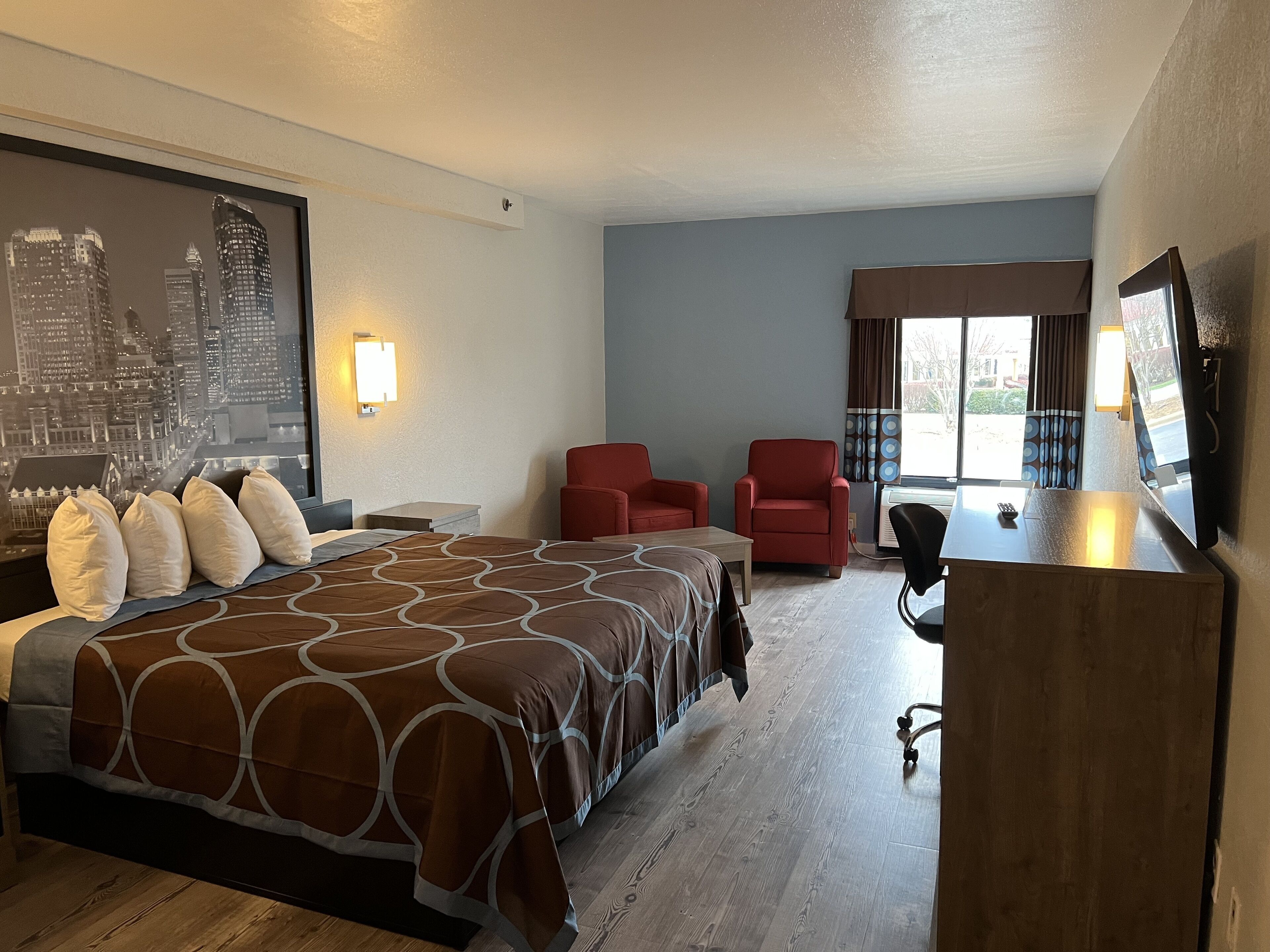Une chambre d’hôtel avec un grand lit, deux fauteuils rouges, un bureau avec une télévision et une vue sur la ville.