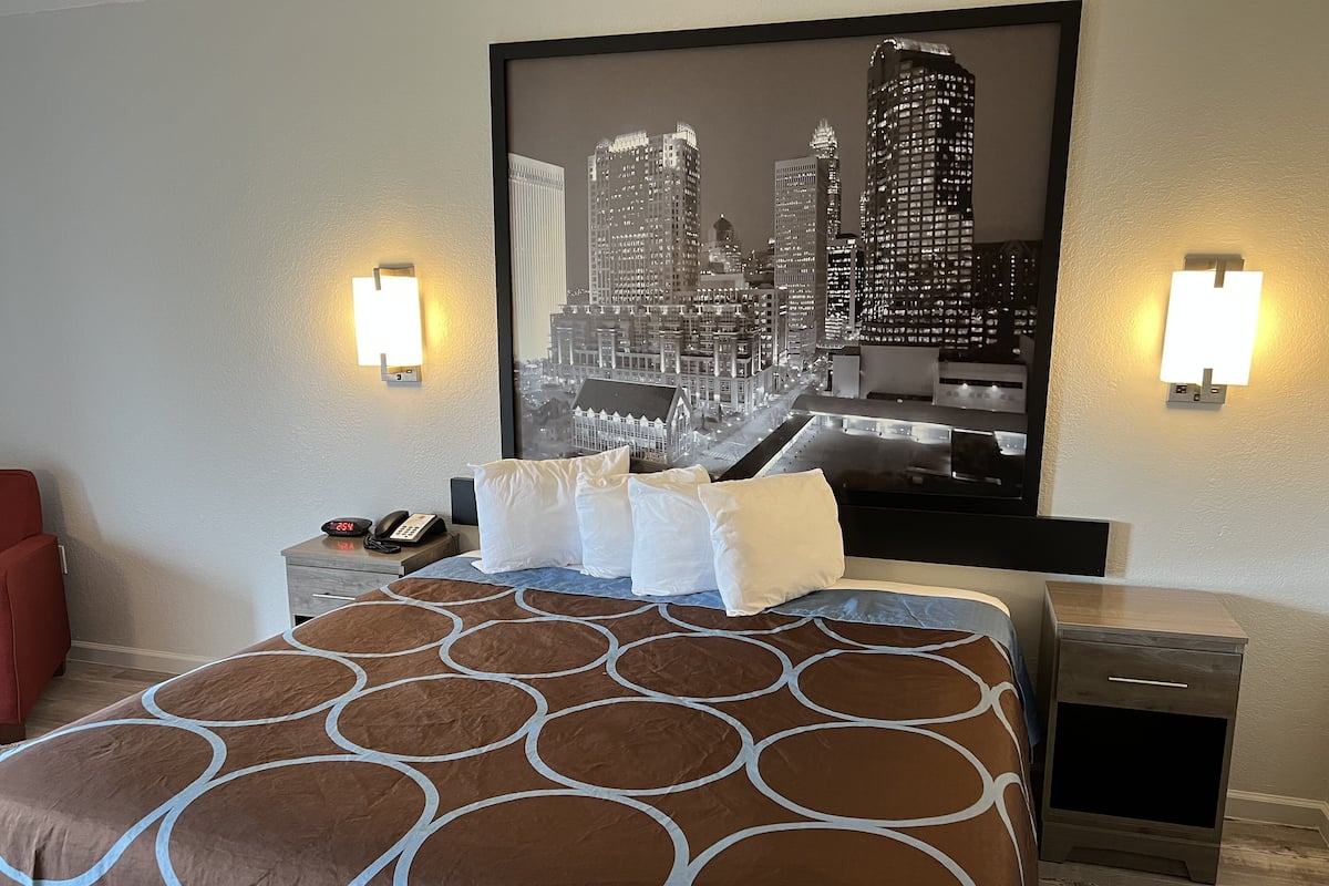 A hotel room with a bed, a nightstand, a red chair, a framed cityscape picture, and wall sconces.