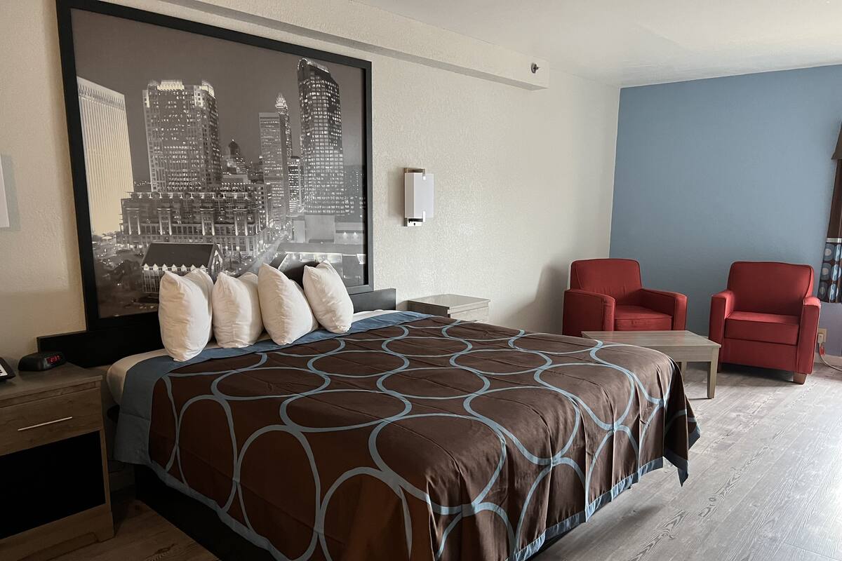 A hotel room with a large bed, a cityscape view, two red chairs, a wooden table, and a nightstand with a clock.