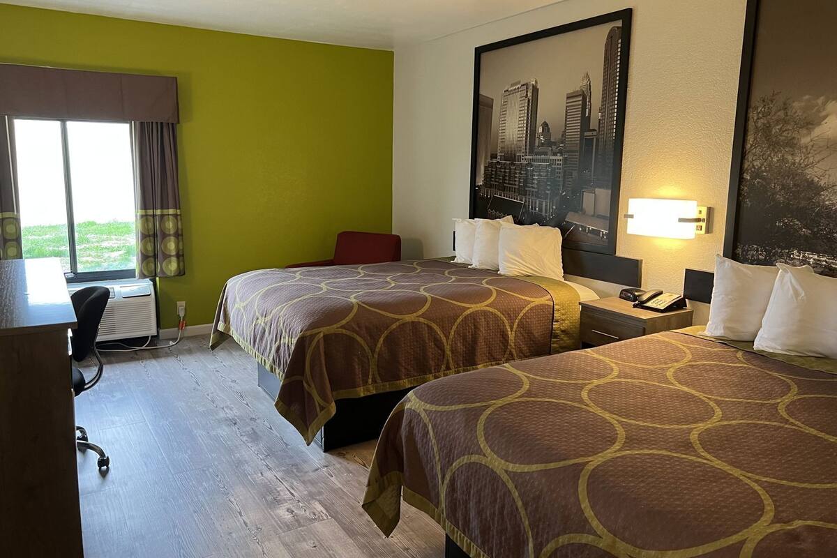 A hotel room with two beds, a desk, a chair, a window with curtains, and a cityscape painting on the wall.