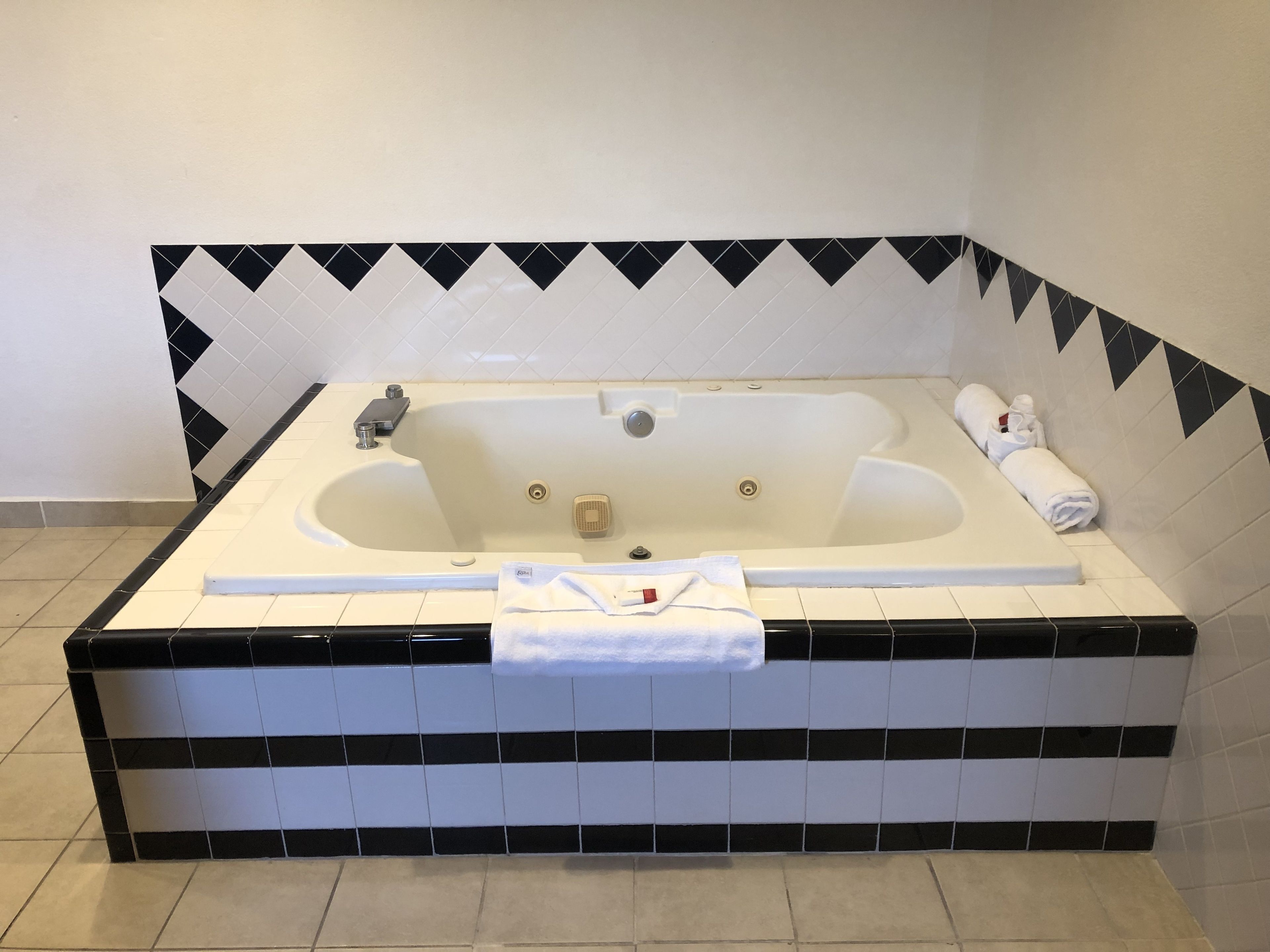 A large, rectangular bathtub with a black and white tile border, a towel, and a bottle on the edge.