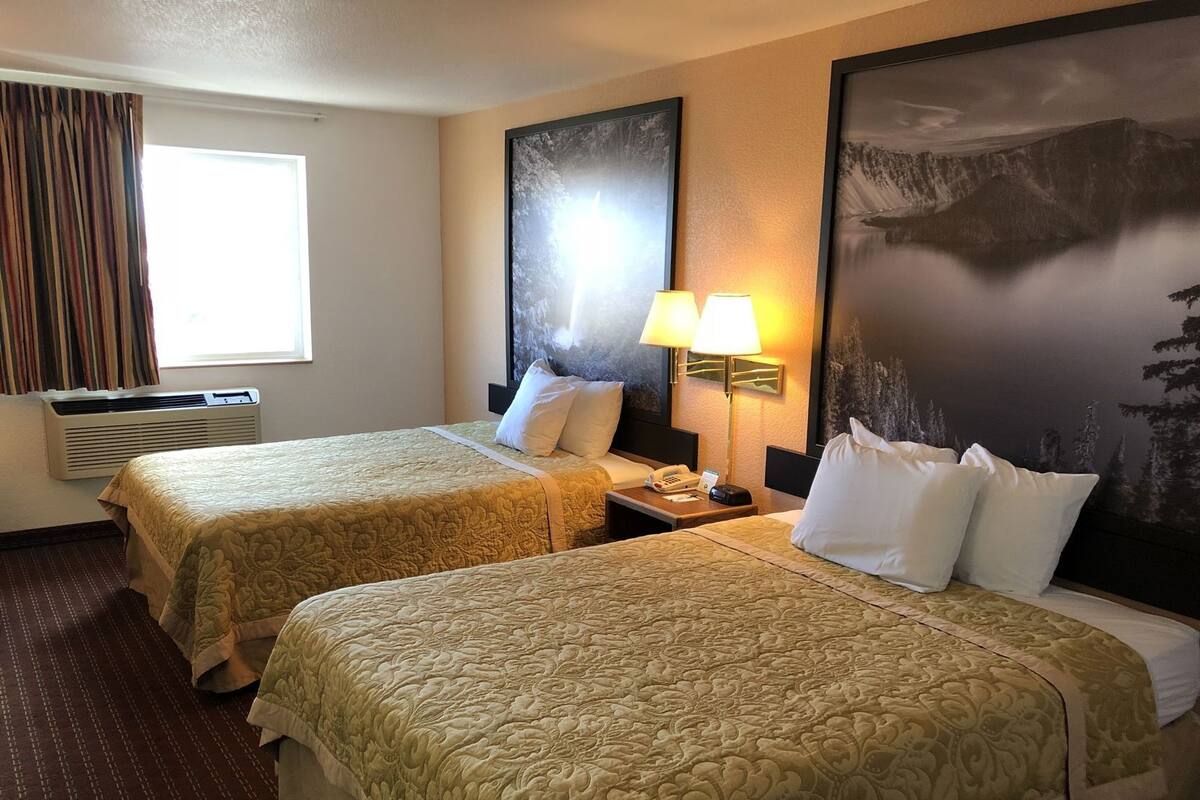 Ein Hotelzimmer mit zwei Betten, einem Fenster mit Vorhängen, einer Klimaanlage, einem Nachttisch mit Lampe und einem großen gerahmten Bild an der Wand.