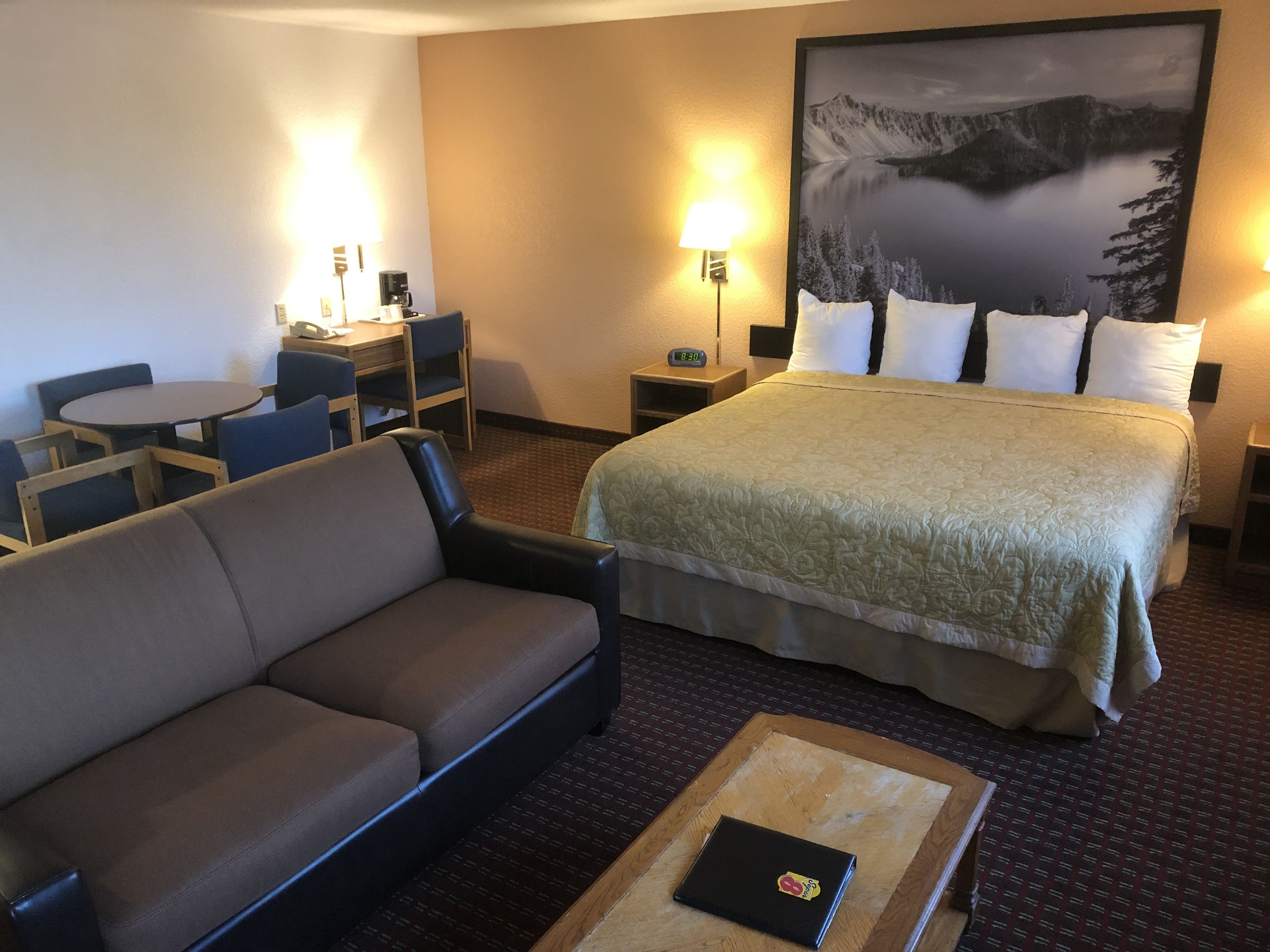 Ein Hotelzimmer mit einem Bett, einem Sofa, einem Schreibtisch mit Stühlen, einer Lampe und einem Gemälde einer Berglandschaft.