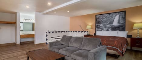 Una habitación de hotel moderna con sofá, una mesa de madera, una cama con cubrecama estampado, una mesita de noche con lámpara y un cuadro grande colgado en la pared con una cascada.