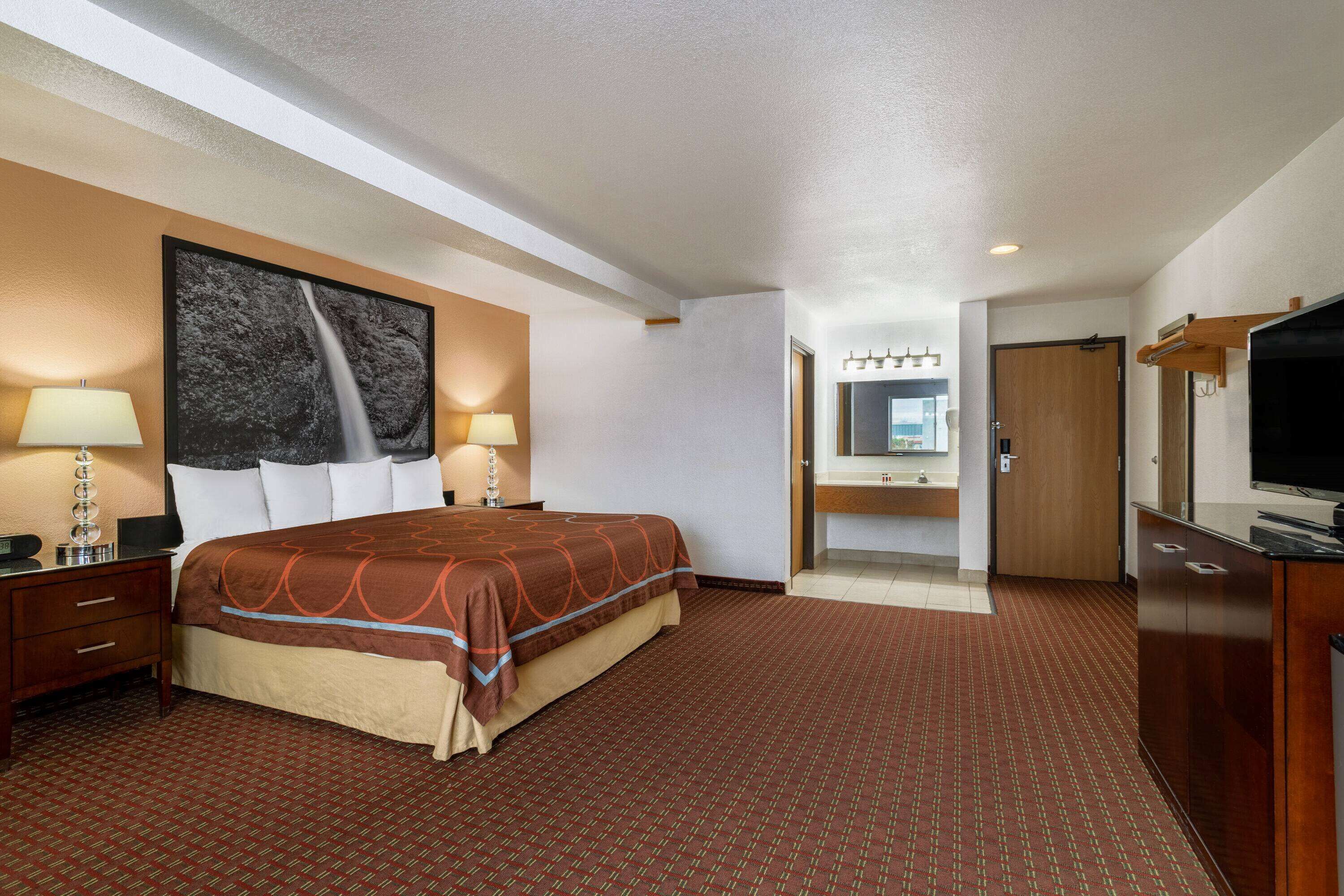 Quarto de hotel com uma cama grande, duas mesas de cabeceira com luminárias, um espelho amplo, televisão e um banheiro visível através de uma porta aberta.