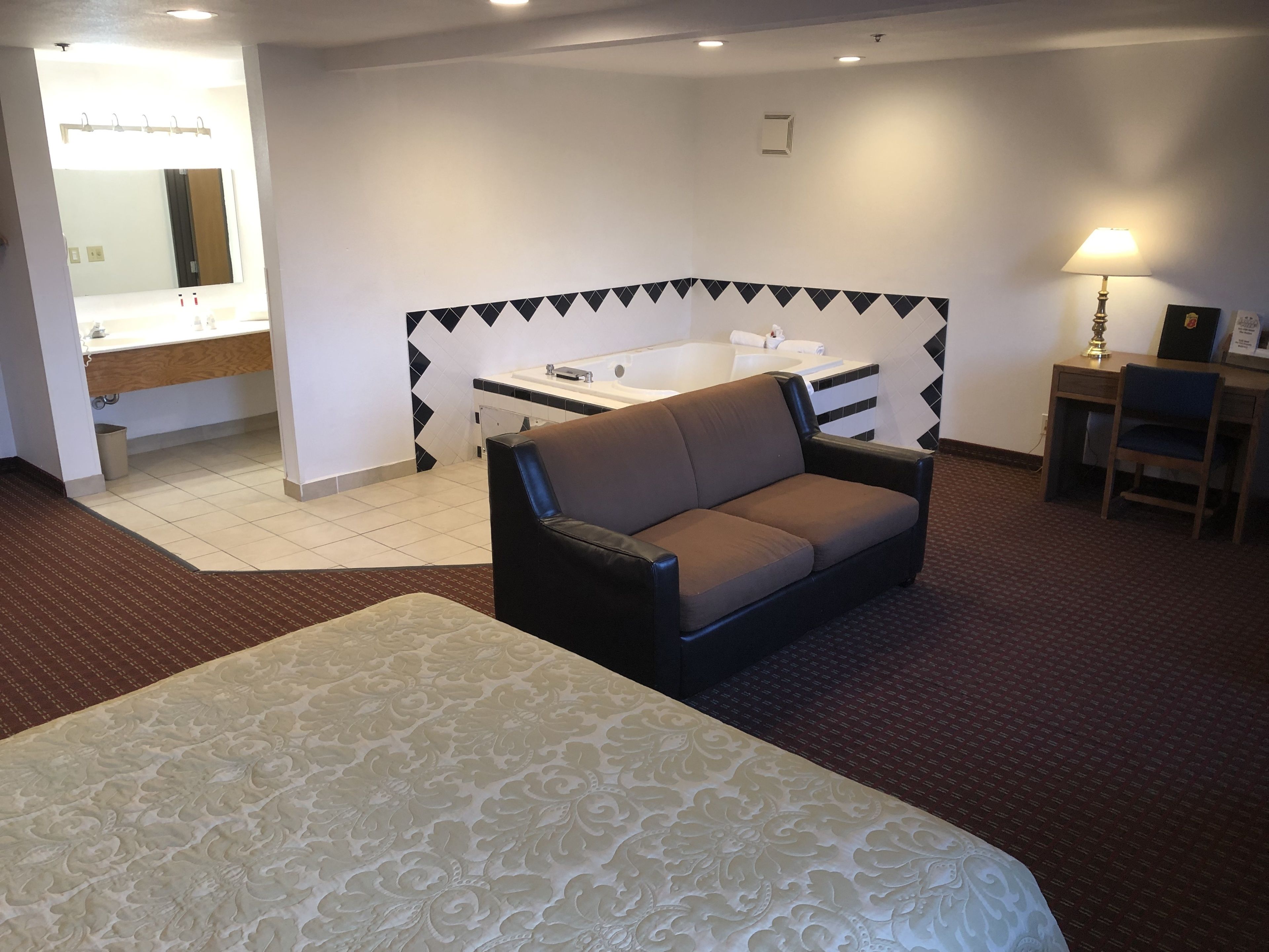 A hotel room with a bed, a sofa, a desk, a chair, a lamp, a bathroom with a sink and a bathtub.