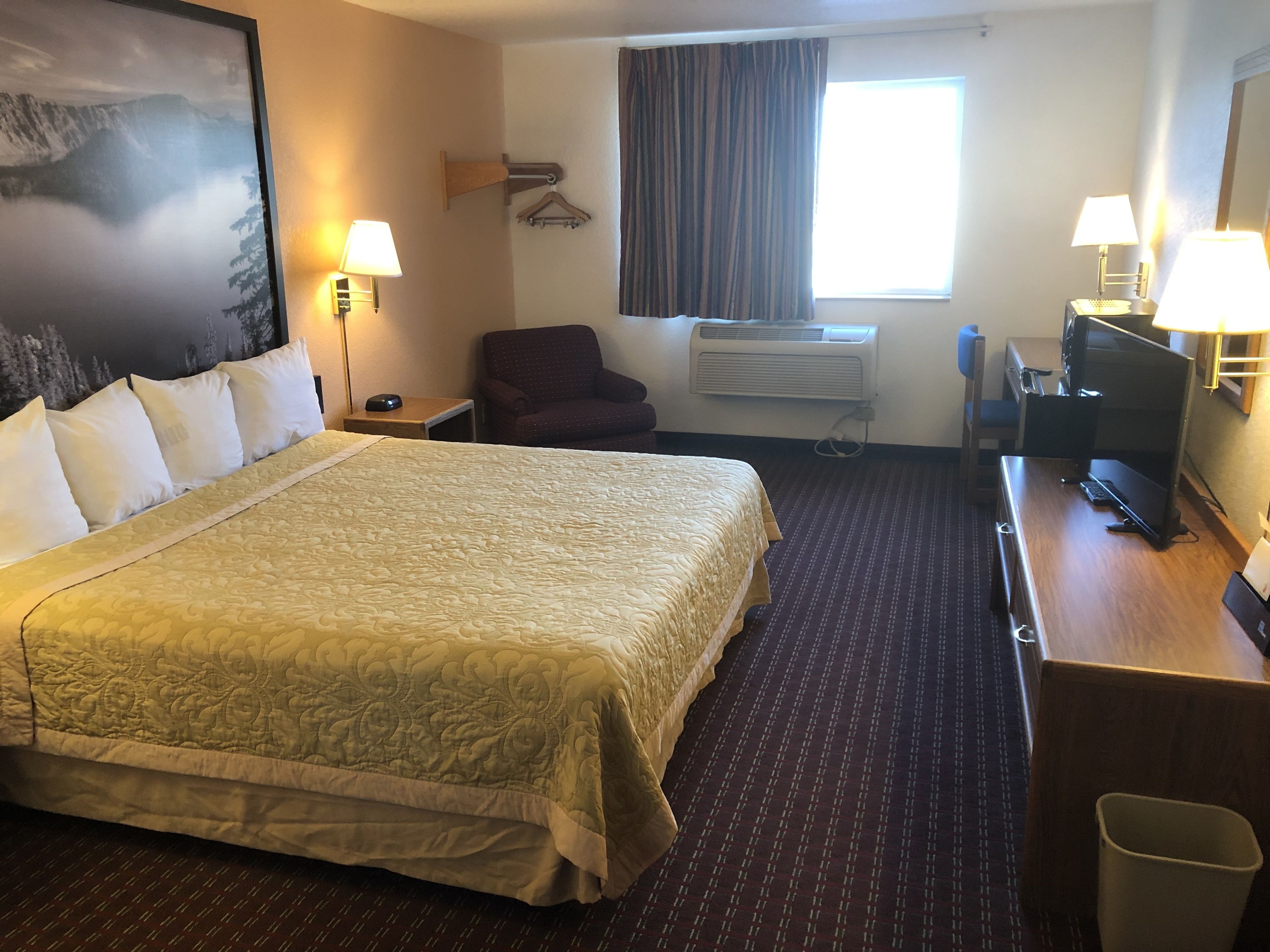 Habitación de hotel con una cama grande, dos lámparas de noche, un escritorio con una computadora, una silla, un equipo de aire acondicionado y una ventana con cortinas.