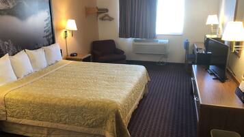 A hotel room with a large bed, two bedside lamps, a desk with a computer, a chair, an air conditioning unit, and a window with curtains.