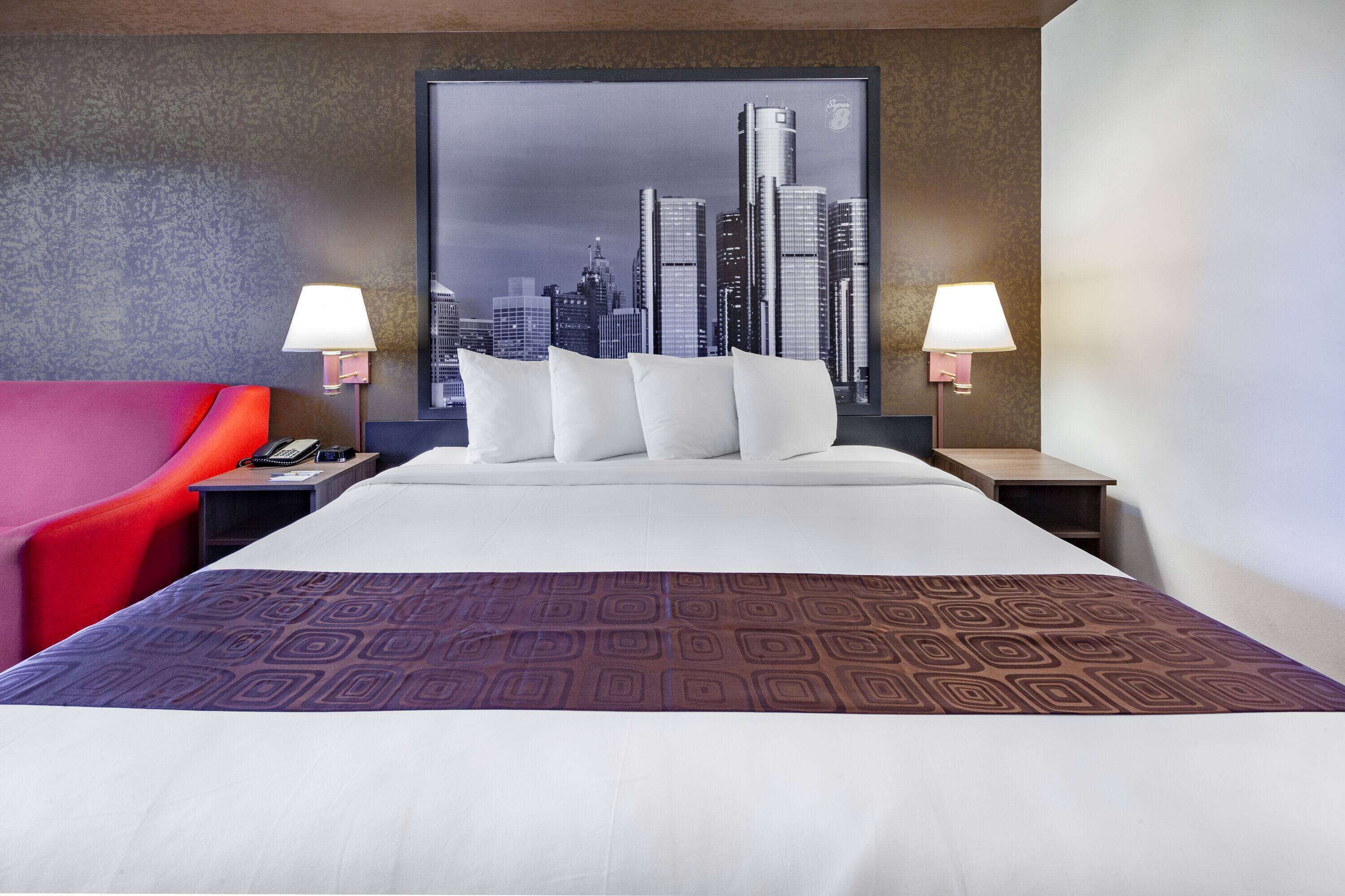 A hotel room with a large bed, two bedside tables, a red chair, and a cityscape view.