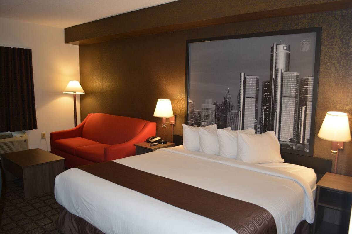 Une chambre d’hôtel avec un lit, un fauteuil rouge, deux lampes de chevet, une décoration murale représentant un paysage urbain et un téléphone sur la table de chevet.