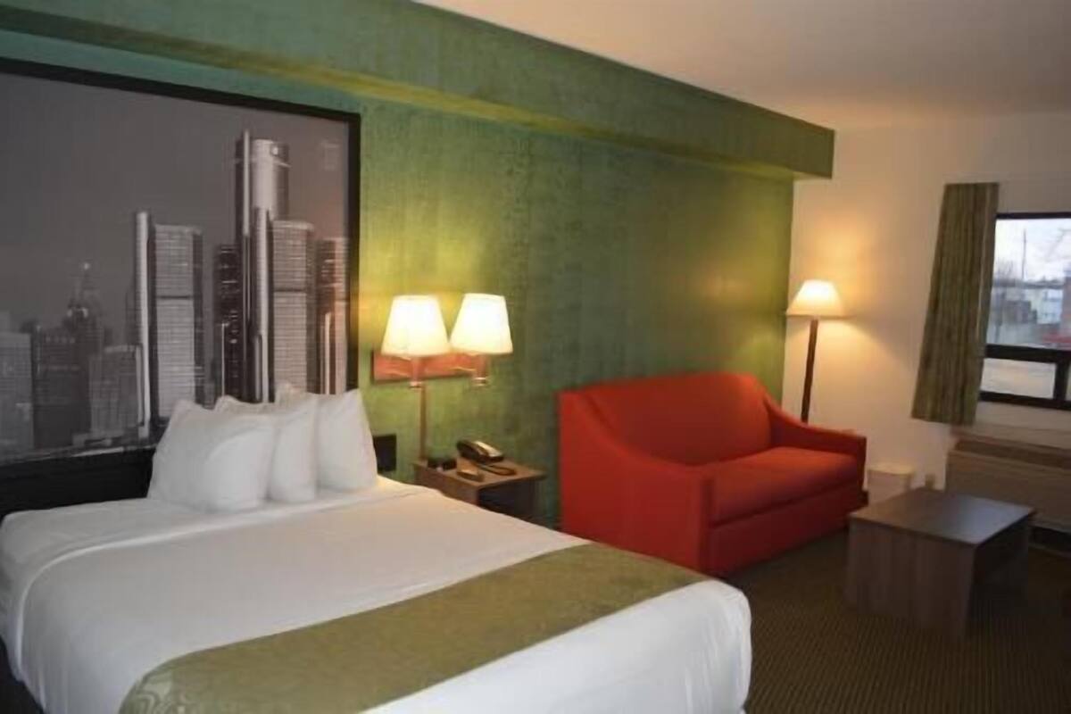 Une chambre d’hôtel avec un lit, un fauteuil rouge, deux lampes, une petite table et un tableau représentant un paysage urbain accroché au mur.