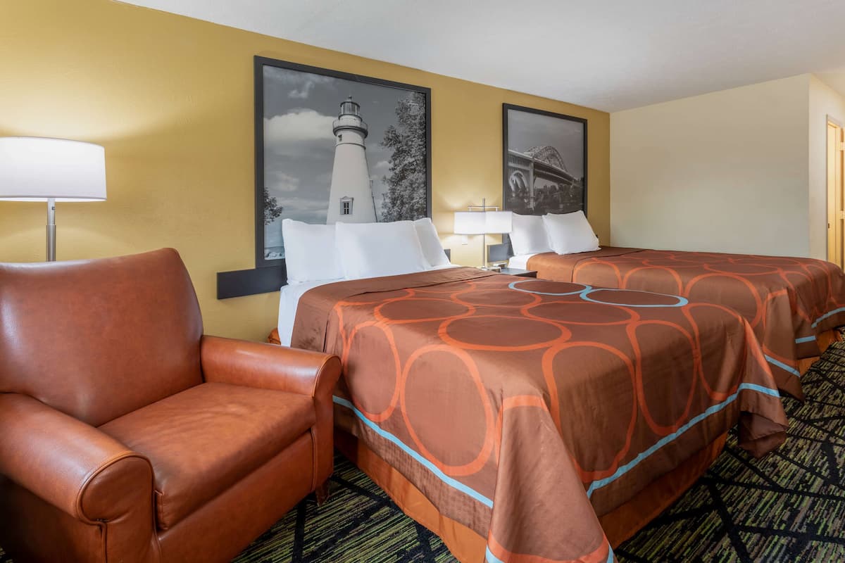 Ein Hotelzimmer mit einem braunen Ledersessel, einem Doppelbett mit orange-blauen Musterbettwäsche, zwei Wandlampen und zwei gerahmten Bildern von Leuchttürmen.