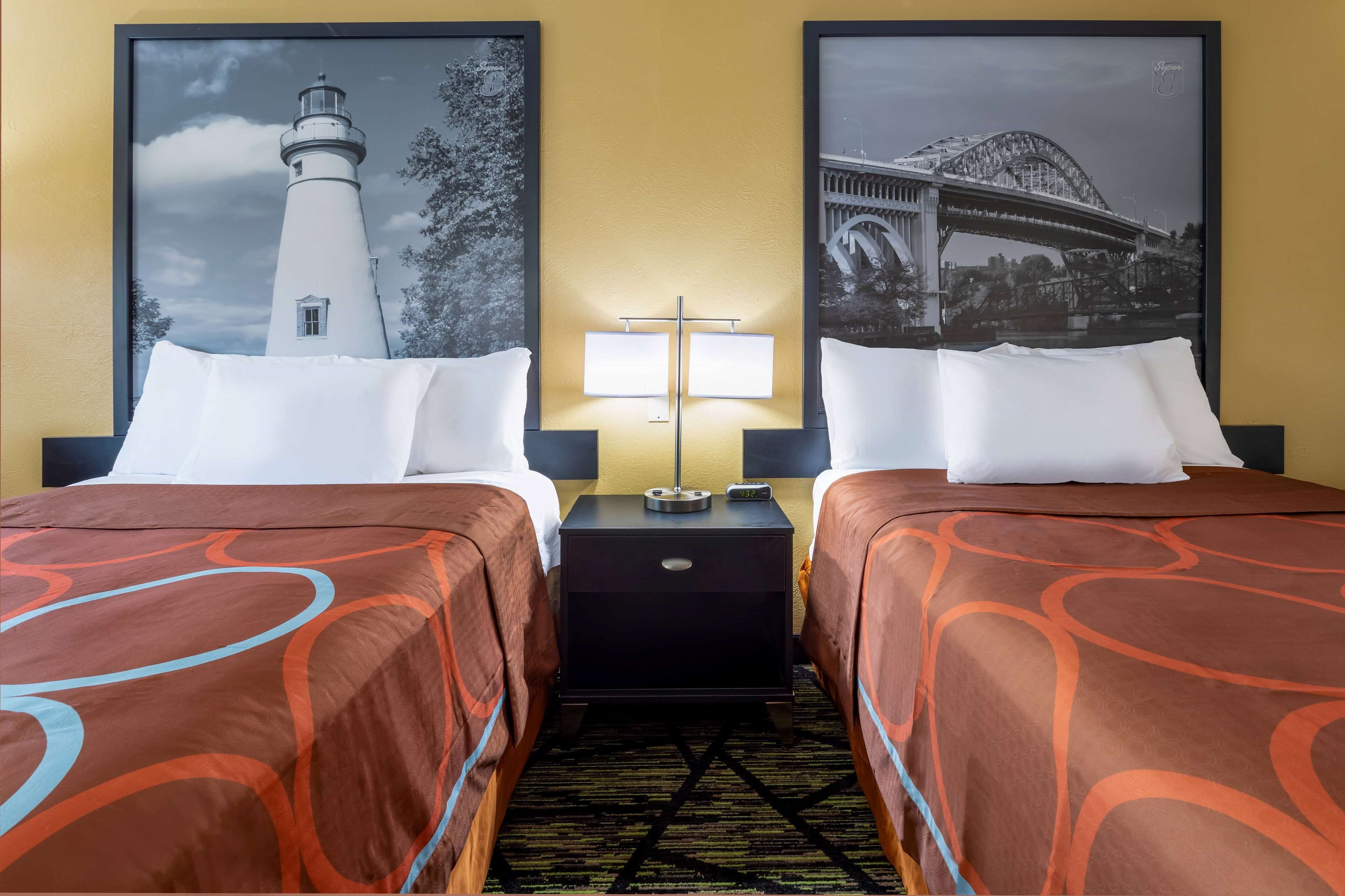 A hotel room with two beds, each with a white pillow and a brown patterned bedspread. There is a nightstand between the beds with a lamp and a clock. The walls are yellow, and there are two framed pictures above the beds, one of a lighthouse and the other of a bridge.