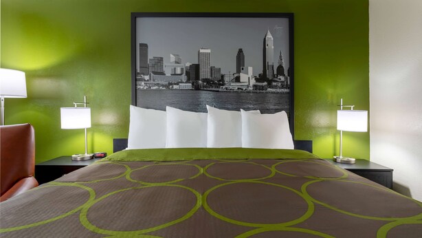 Une chambre d’hôtel avec un mur vert, un grand lit recouvert d’une couvre-lit à motifs marron et vert, deux tables de chevet avec des lampes, et un tableau représentant un paysage urbain accroché au mur.