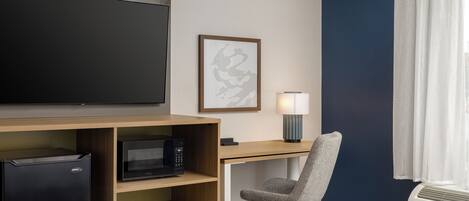 A hotel room with a bed, a desk with a computer, a microwave, a refrigerator, a chair, a lamp, and a large TV on the wall.