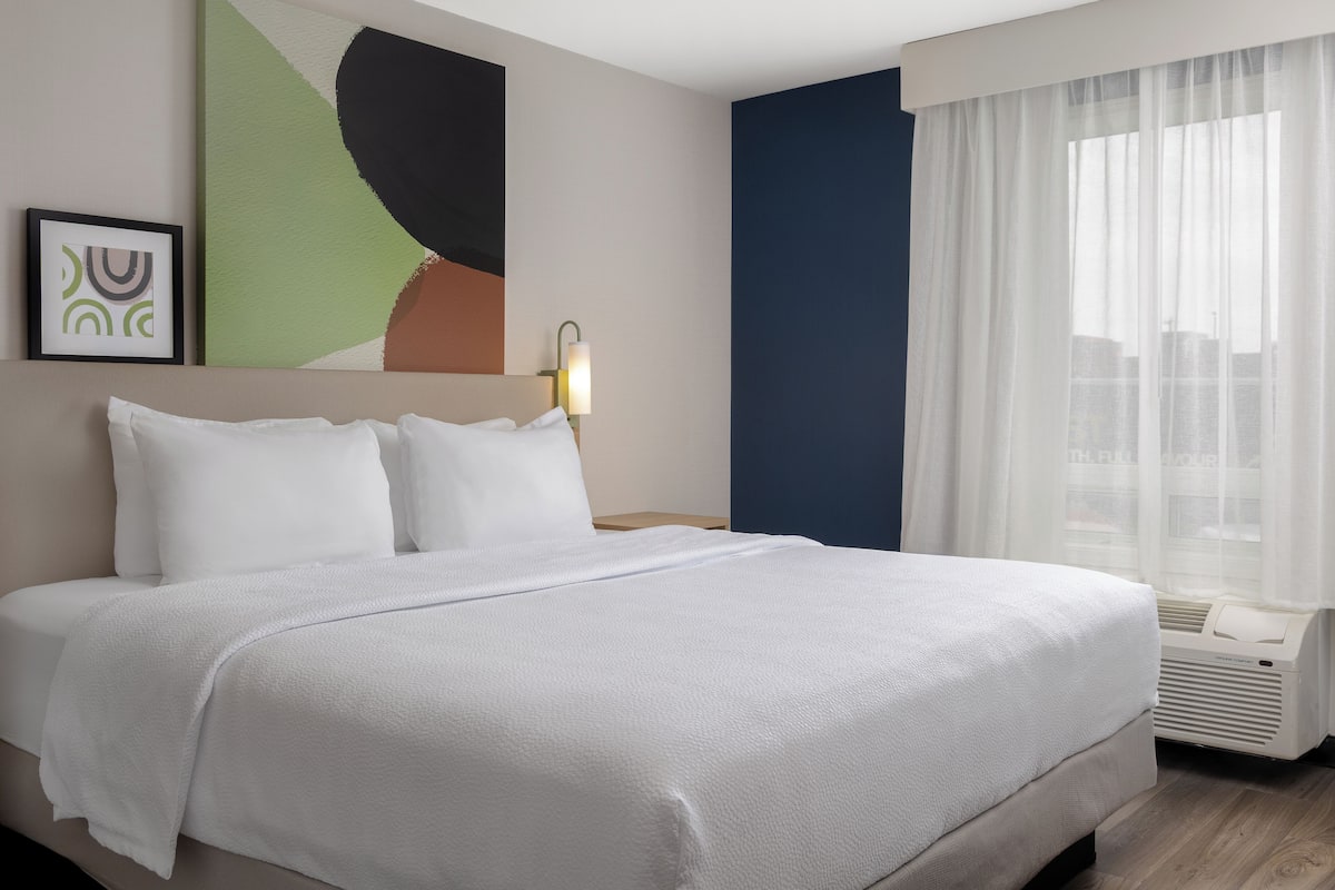 Una cama bien hecha con sábanas blancas en una habitación de hotel con vistas a la ciudad a través de cortinas traslúcidas.