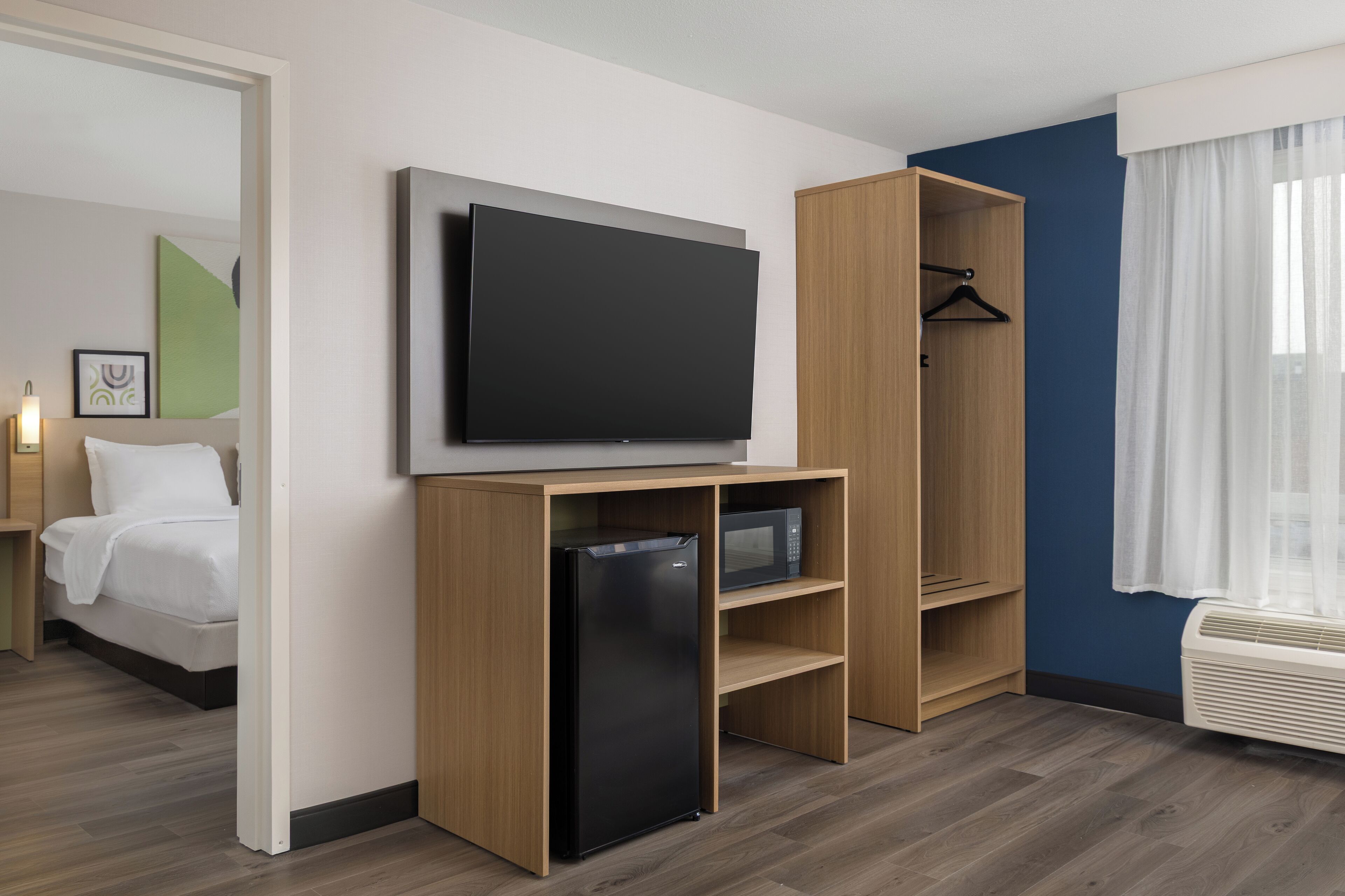 A modern hotel room with a flat-screen TV, a mini-fridge, a wooden wardrobe, and a bed.