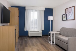 Quarto de hotel moderno com televisão de tela plana, sofá cinza, mesa pequena branca, abajur e uma janela com cortinas translúcidas.
