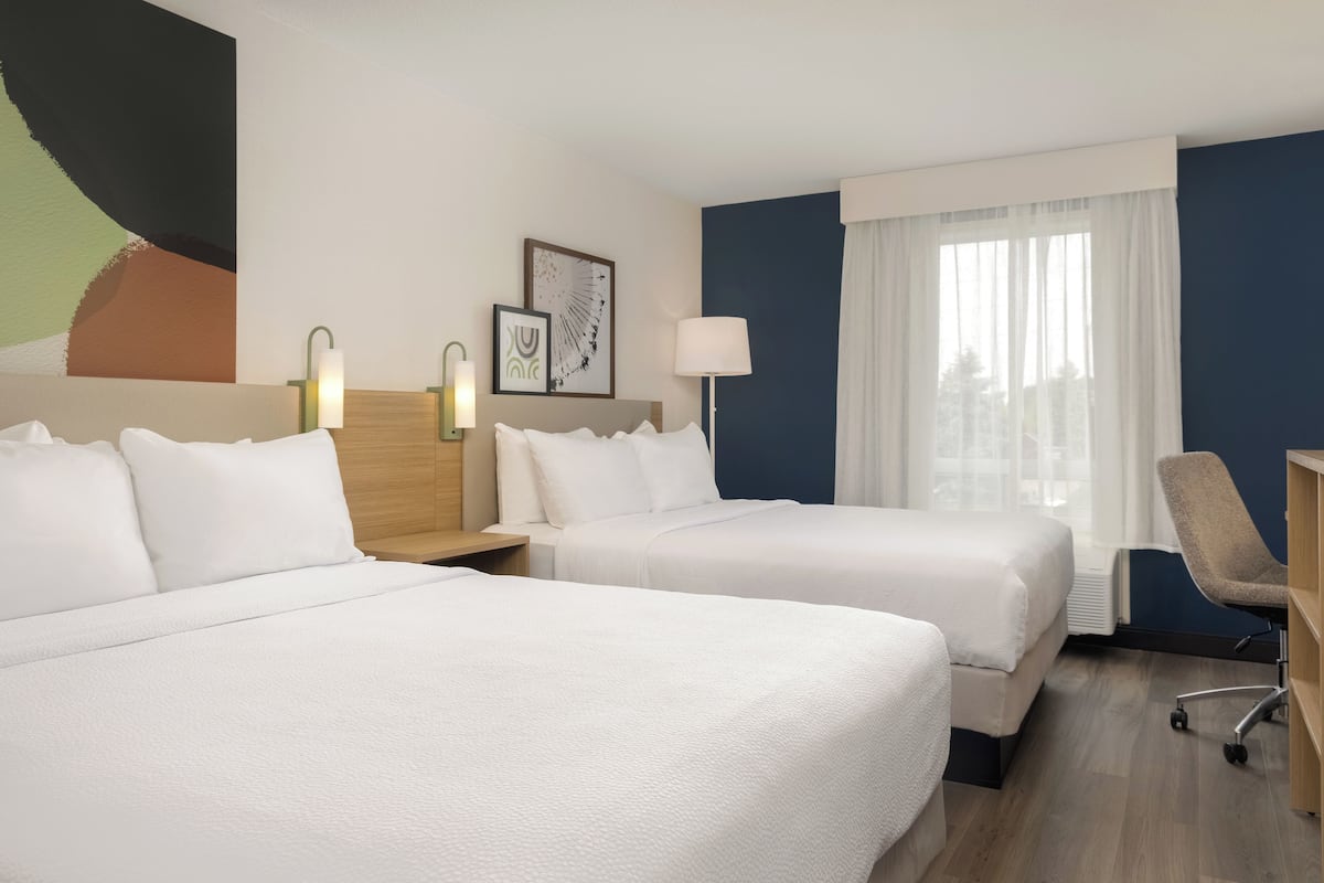Habitación de hotel con dos camas, un escritorio, una silla y un ventanal grande con cortinas.