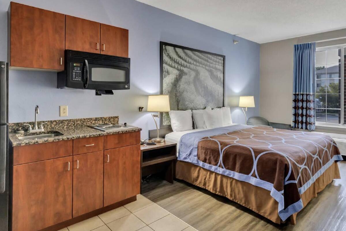 Habitación de hotel con kitchenette, cama con cubrecama estampado, un cuadro en la pared y ventana con cortinas.