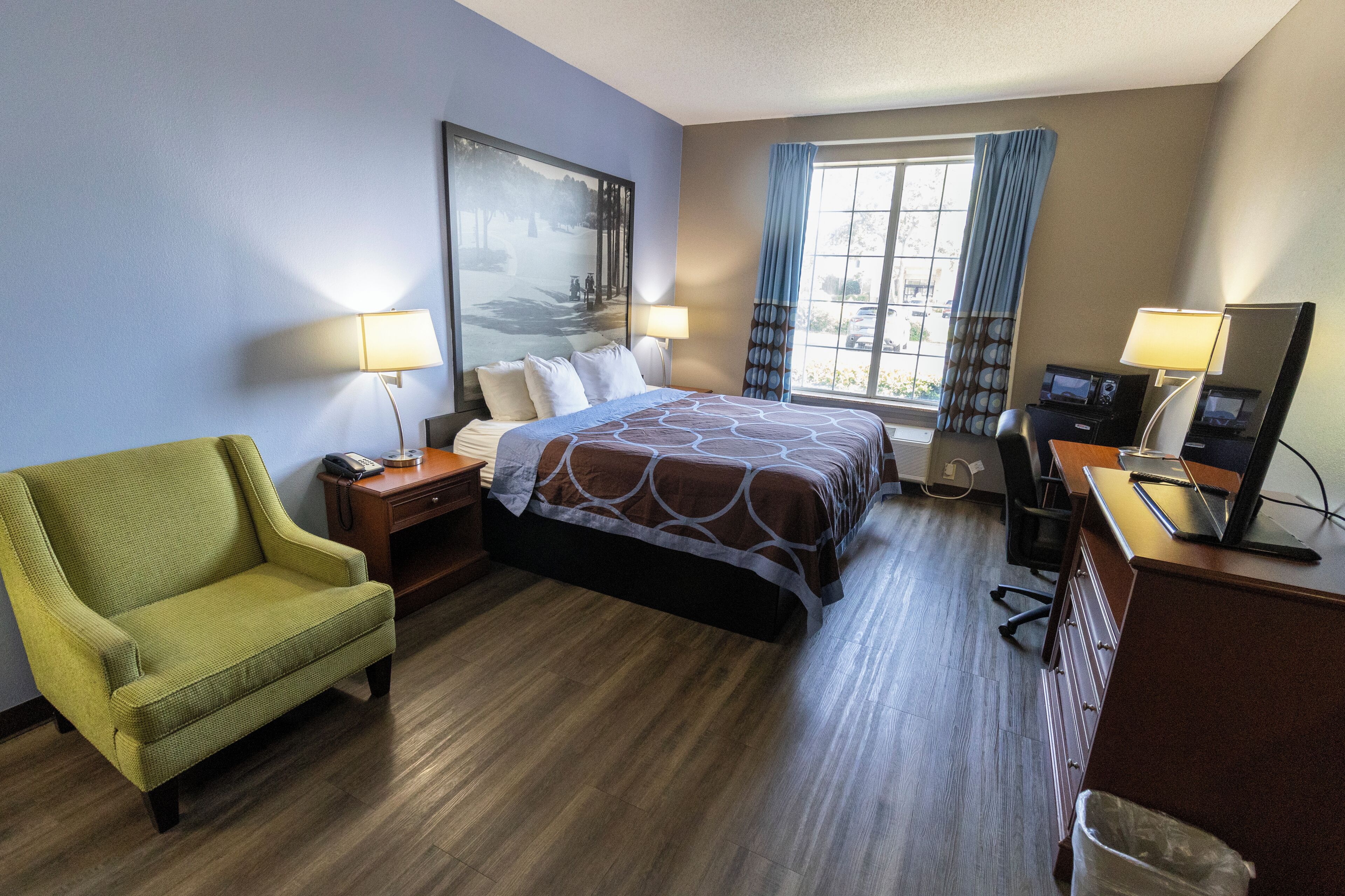Habitación de hotel con cama, escritorio con computadora, silla, lámpara de escritorio y ventana con cortinas.
