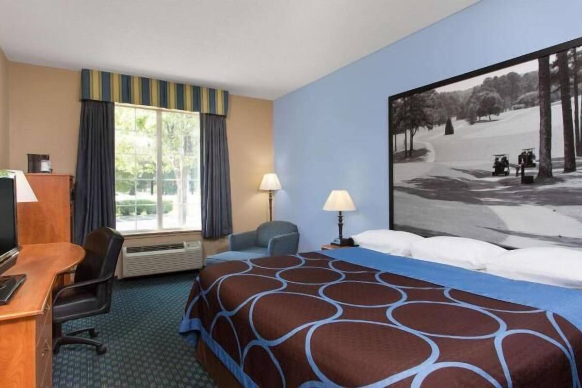 Habitación de hotel con cama, escritorio, silla, televisor y una ventana grande con cortinas.