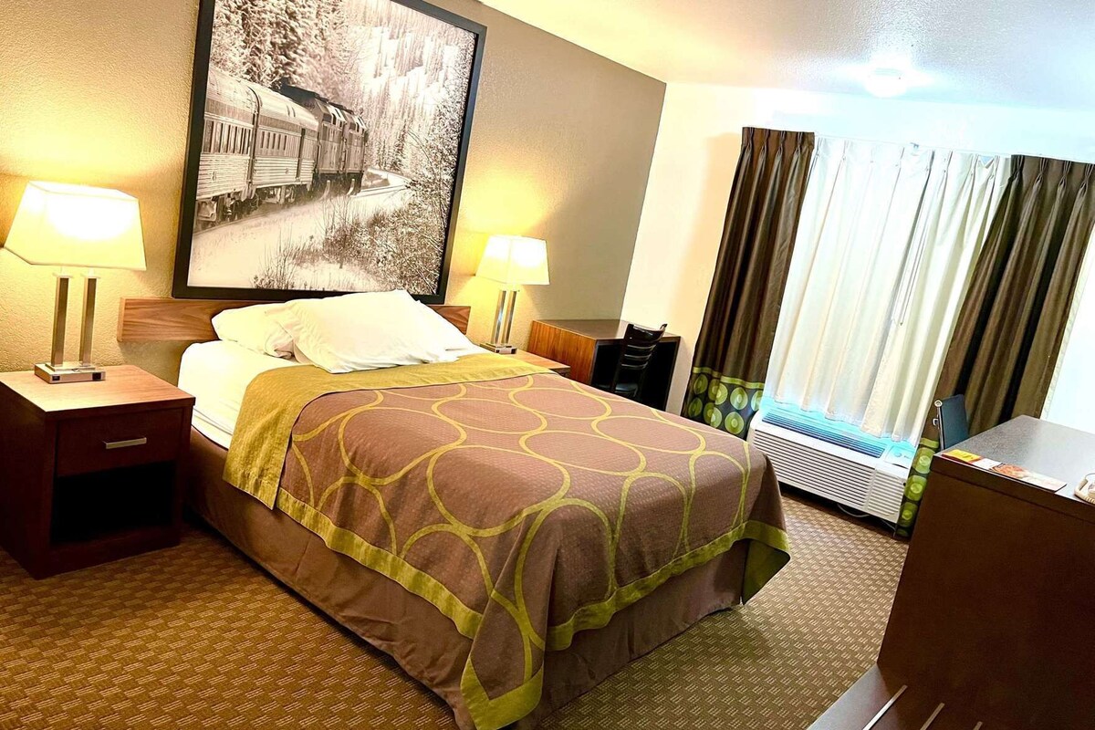 A hotel room with a bed, two bedside tables, a desk, a chair, a large window with curtains, and a wall-mounted air conditioning unit.