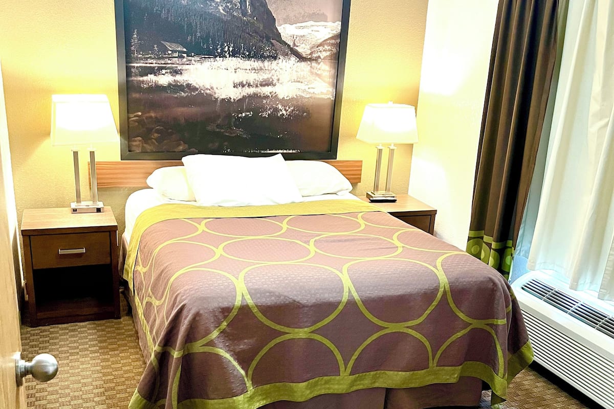Una camera d'albergo con un letto, due abat-jour, un'ampia foto incorniciata di un paesaggio montano e una finestra con tende.