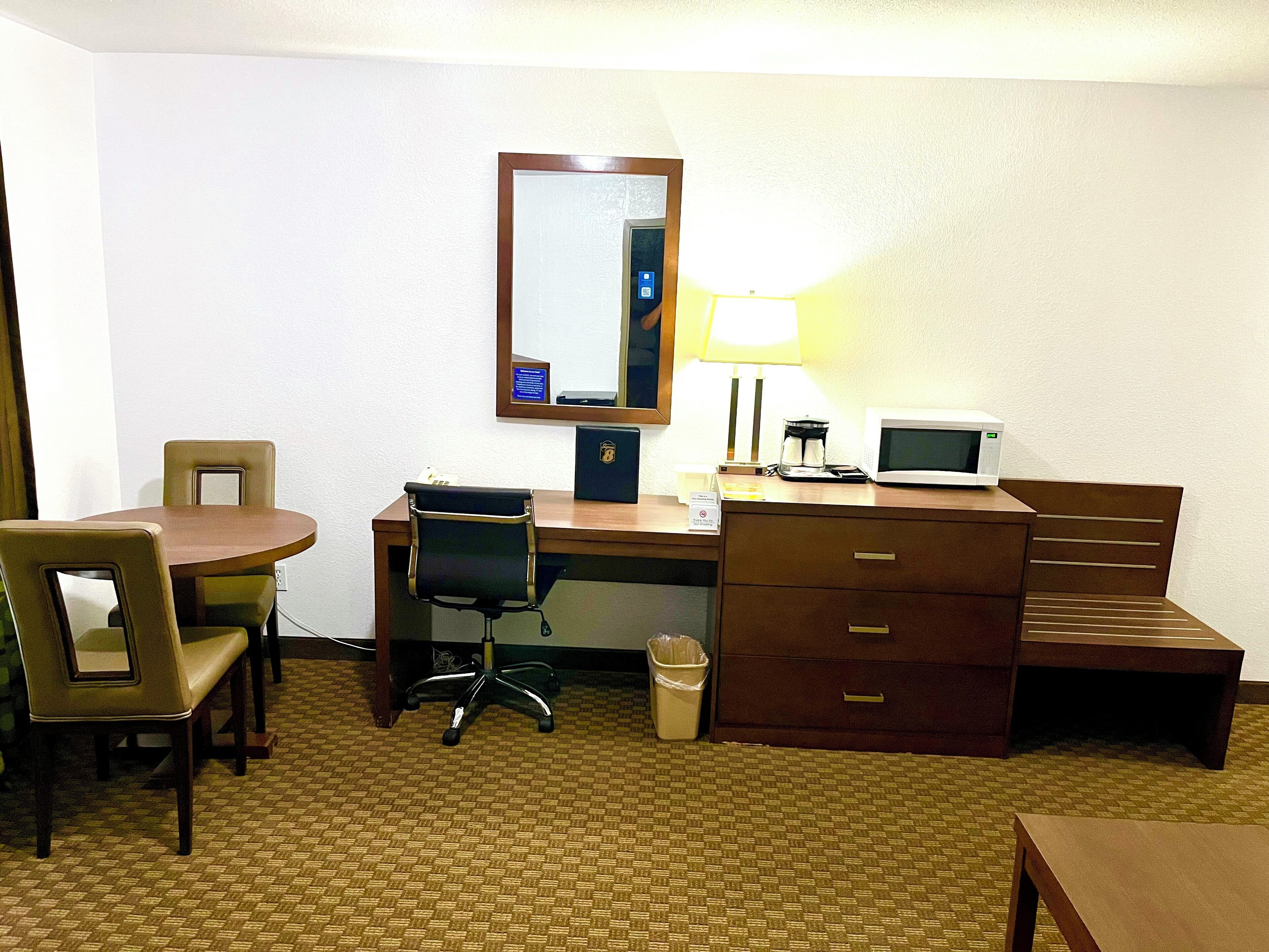 A hotel room with a desk, two chairs, a lamp, a microwave, a trash can, and a mirror.