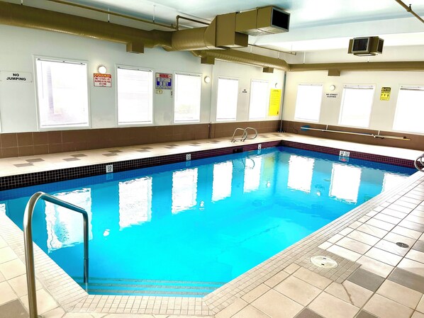 An indoor swimming pool with clear water, a tiled deck, and multiple doors with various signs. - Super 8 by Wyndham Hinton (Hinton)