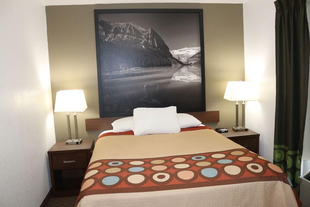 Una camera d'albergo con un letto, due lampade da comodino, un comodino, un quadro grande appeso alla parete e una finestra con tende.