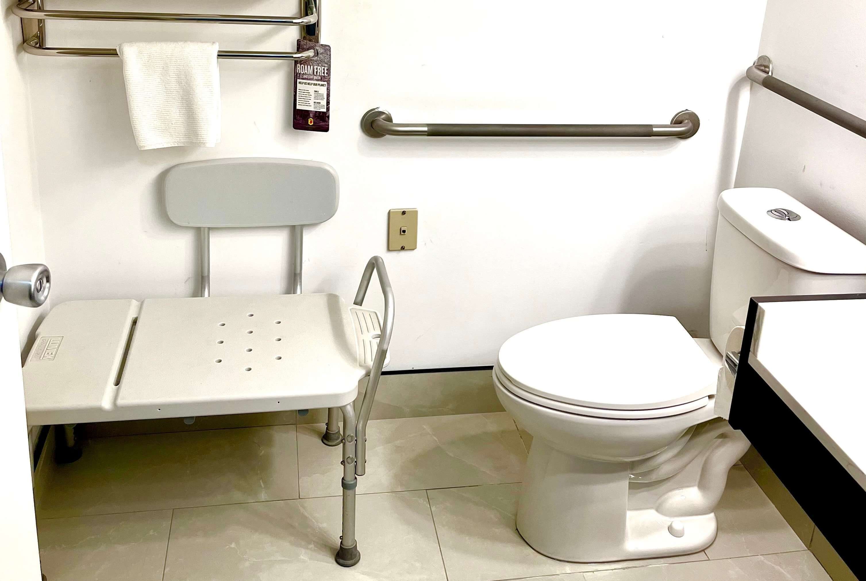 Un baño con inodoro blanco, barra de apoyo, lavabo blanco y asiento de ducha blanco.
