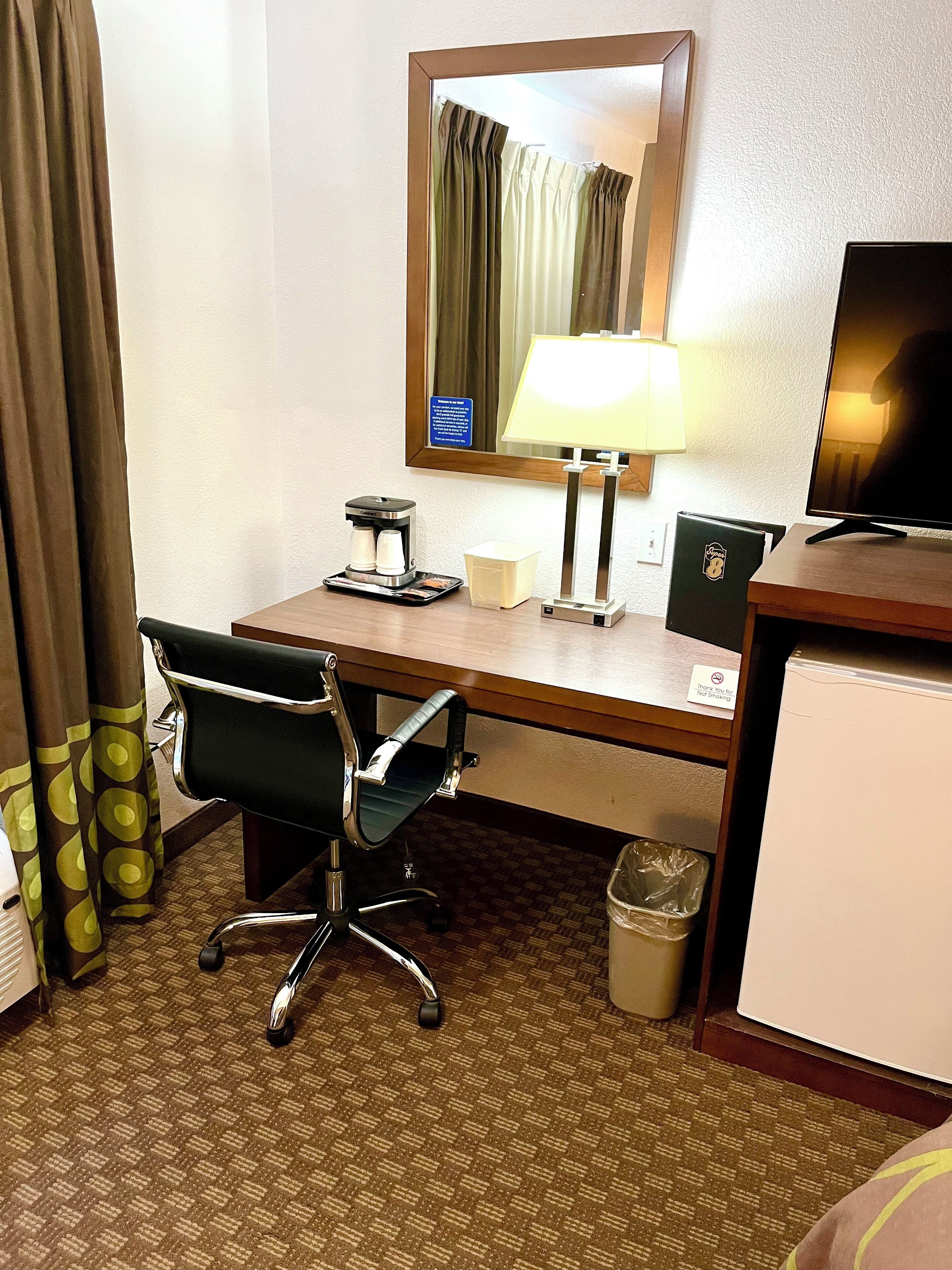 A hotel room with a wooden desk, a chair, a coffee maker, a lamp, a television, and a trash bin.