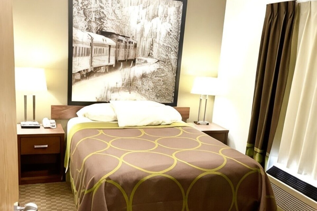 A hotel room with a bed, two bedside tables, a nightstand, a large framed picture on the wall, and a window with curtains.