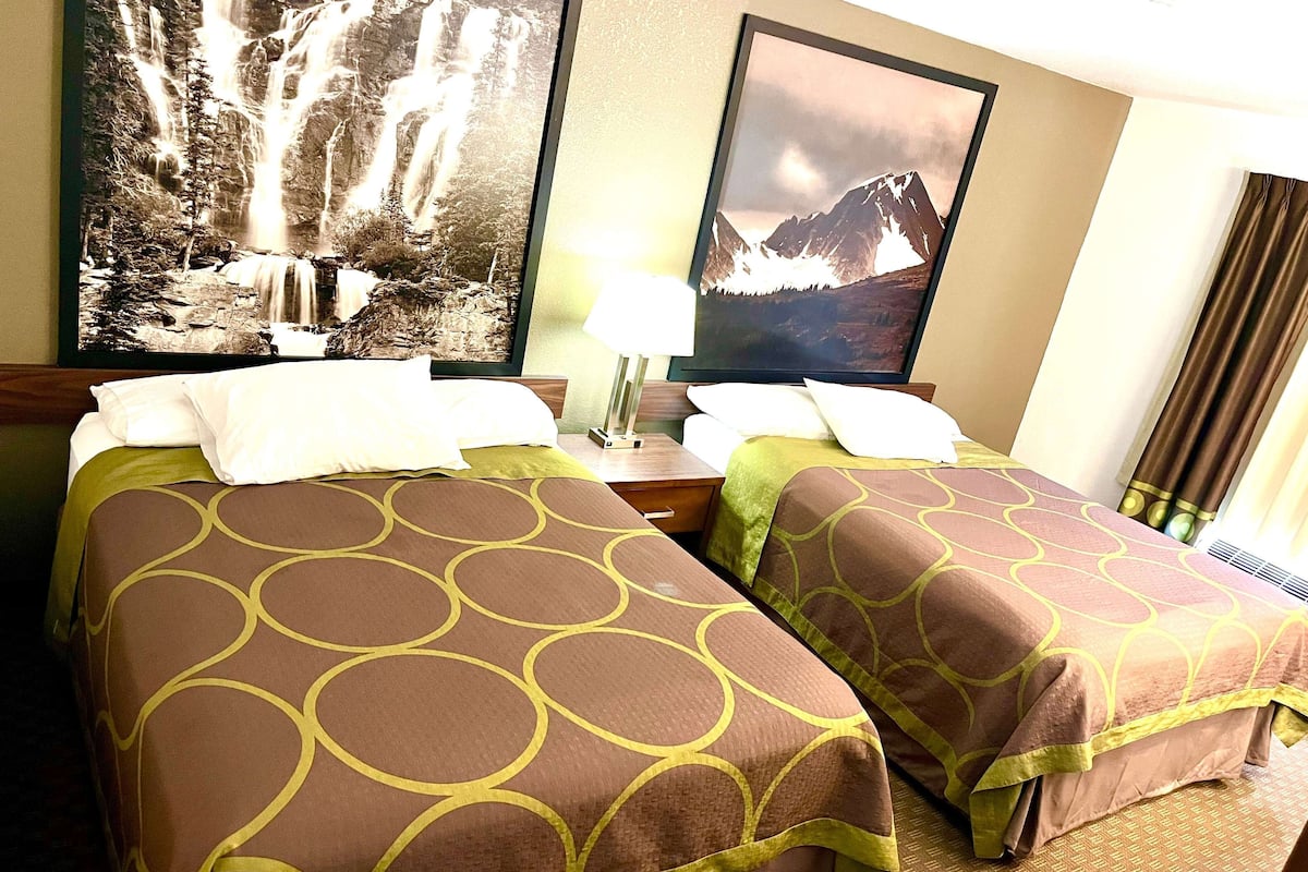 Una camera d'albergo con due letti, ognuno con una coperta a motivi marroni e verdi, un comodino con una lampada e due quadri appesi raffiguranti cascate e montagne.