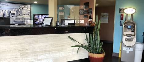 A hotel reception area with a wooden reception desk, a potted plant, a house phone, and an ATM.