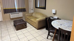Une chambre d’hôtel avec un canapé, une petite table recouverte d’une nappe à motifs, une chaise, un banc, un climatiseur fixé au mur, une porte et une fenêtre avec des rideaux.