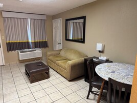 Habitación de hotel con sofá, una pequeña mesa con mantel estampado, una silla, un banco, aire acondicionado instalado en la pared, una puerta y una ventana con cortinas.