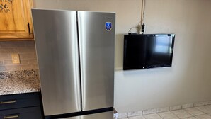 Un réfrigérateur en acier inoxydable avec un autocollant indiquant une garantie de deux ans, un téléviseur noir fixé au mur et un carrelage au sol dans la cuisine.