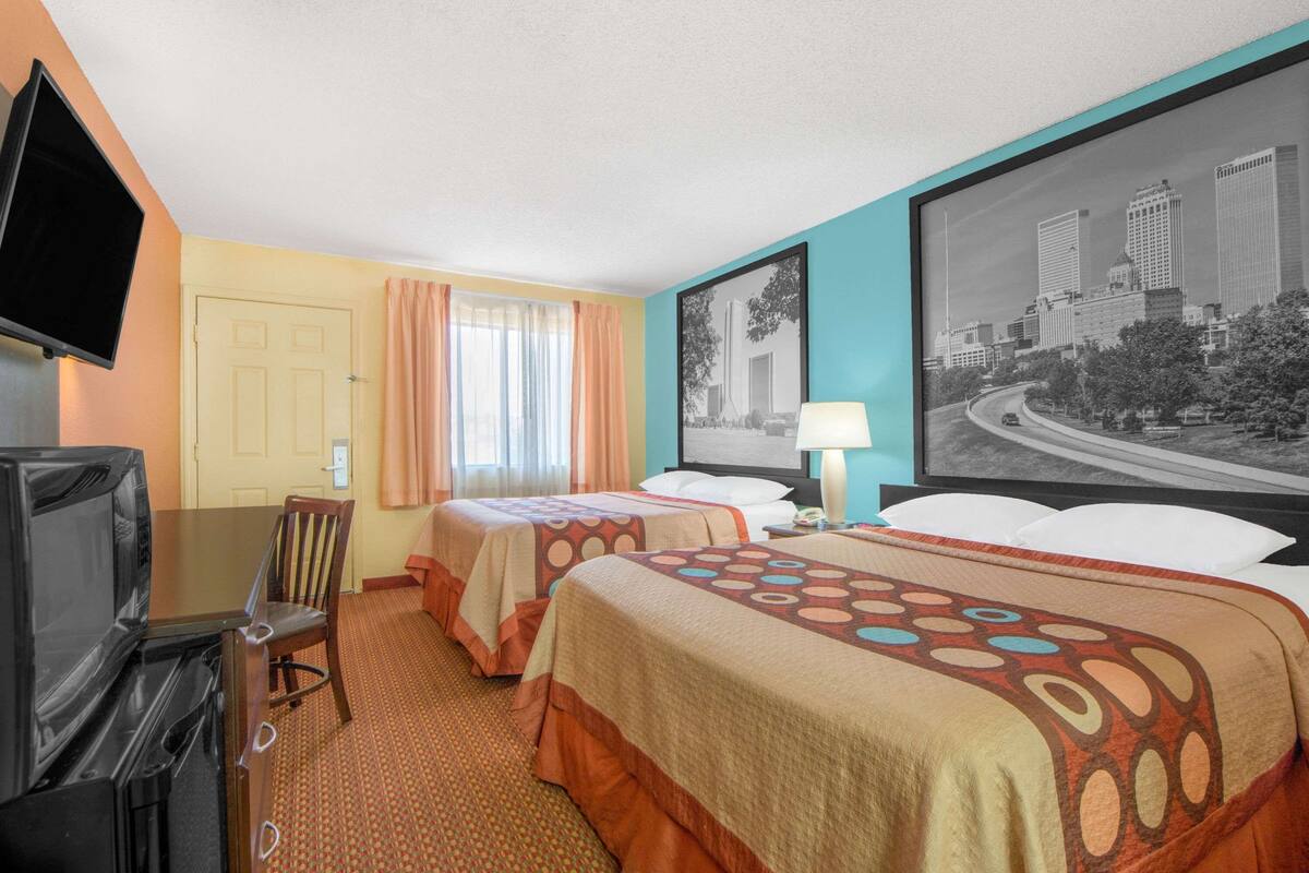 Una camera d'albergo con due letti, una televisione, una scrivania con una sedia, una finestra con tende e un quadro raffigurante un panorama urbano.