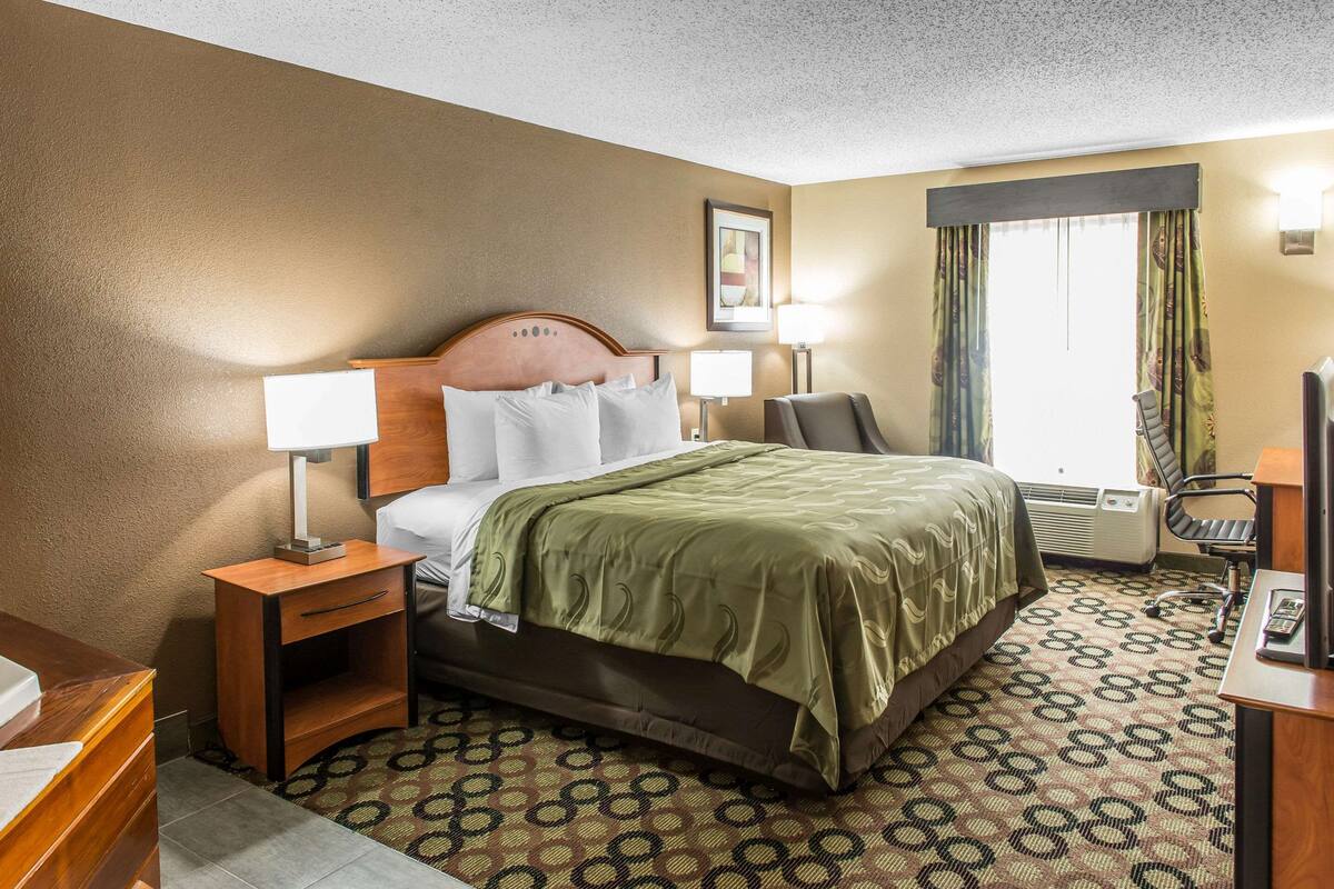 A hotel room with a bed, two bedside tables with lamps, a desk with a chair, and a window with curtains.