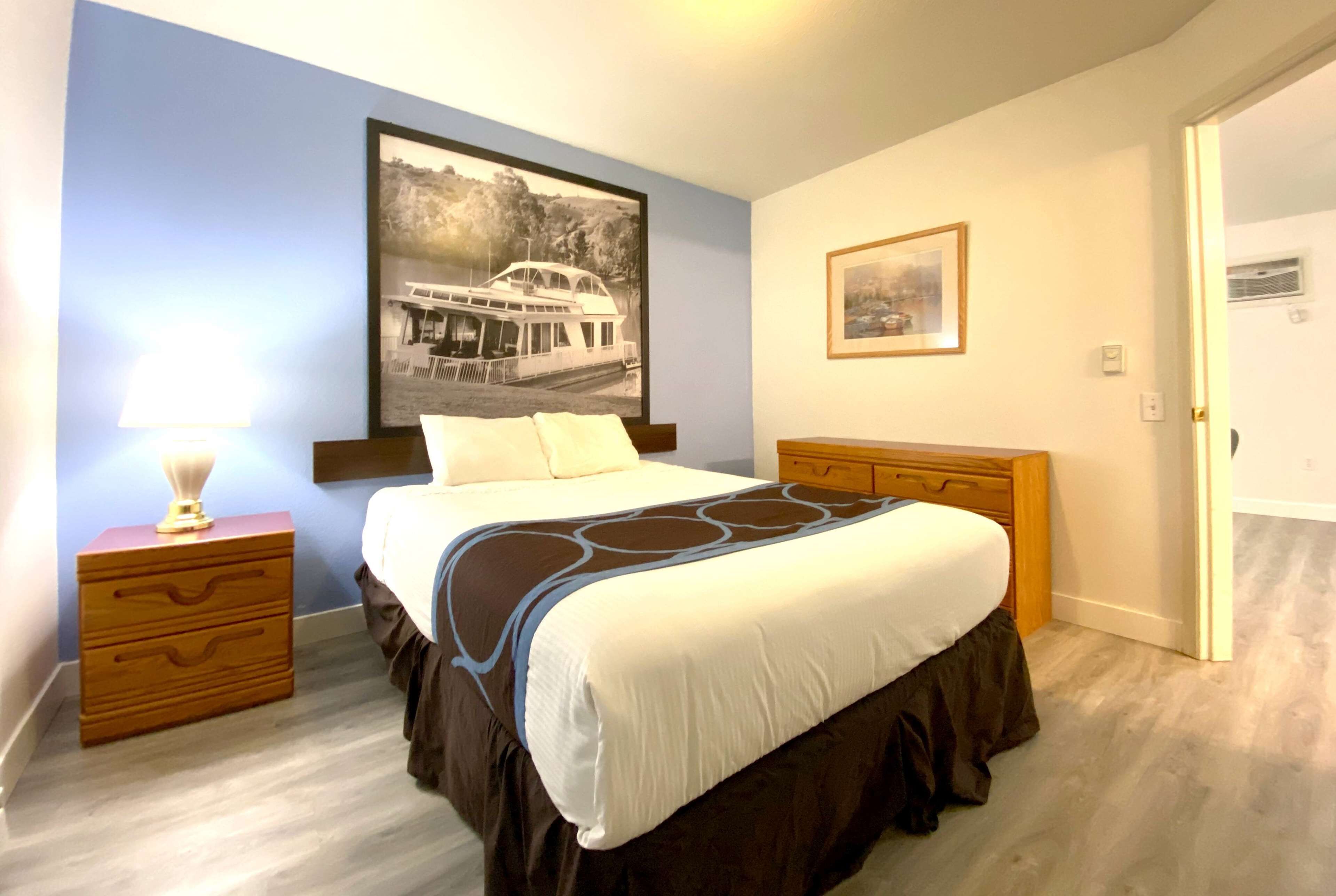 Une chambre à coucher comprenant un lit, une table de chevet, une commode en bois, un tableau encadré accroché au mur et une porte.
