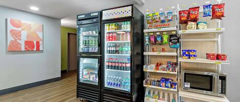 Un hall de hotel con dos refrigeradores de bebidas llenos de diferentes opciones, un microondas en un estante y dispensadores de snacks.