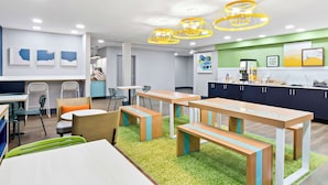 A modern dining area with wooden tables and benches, a kitchen in the background, and artwork on the walls. - Spark by Hilton Winston-Salem University (Winston-Salem)