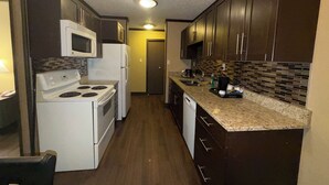 A compact kitchen with a stove, microwave, and refrigerator. It features dark wooden cabinets and a tiled backsplash. - Siding 16 Lodge (Wetaskiwin)