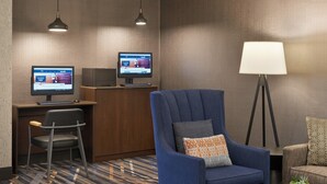 Habitación de hotel con dos monitores de computadora, un sillón azul, una mesita con una planta y una lámpara de pie.