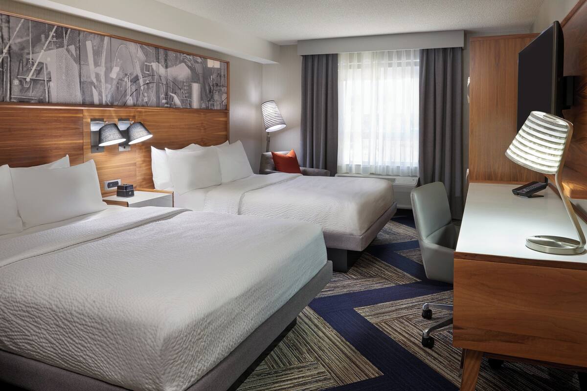 A hotel room with two beds, a desk, a chair, a TV, and a large window with curtains.