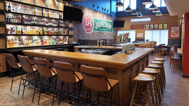 Zona de bar con taburetes, una barra y estantes llenos de botellas de licores.