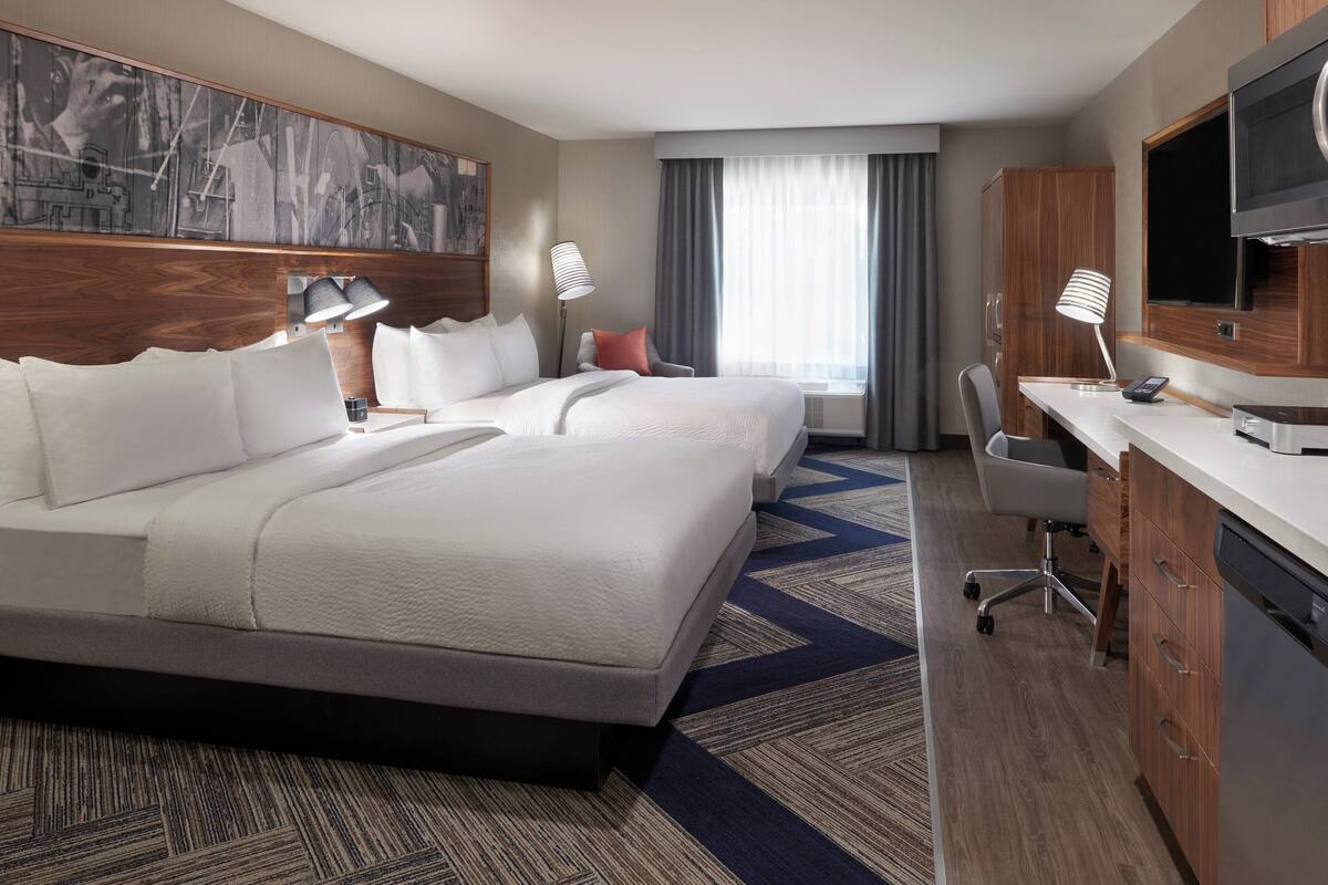 A hotel room with two large beds, a desk, a chair, a television, and a large window with curtains.