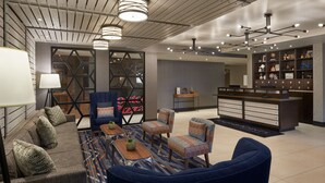 Un moderno vestíbulo de hotel con un sofá, sillones, una mesa de centro, una recepción y una pared con estanterías que exhiben diversos objetos.